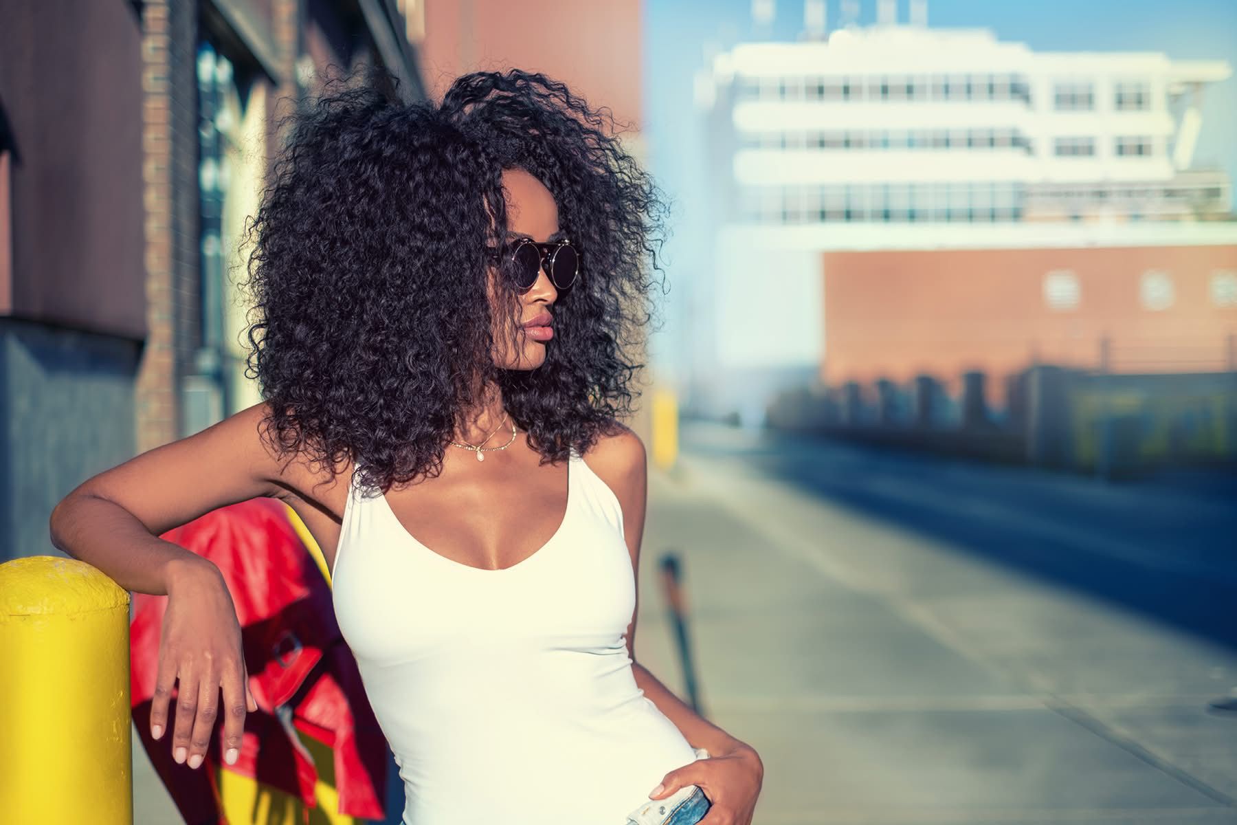 Mimi in a white tank top leaning on a yellow bollard at golden hour