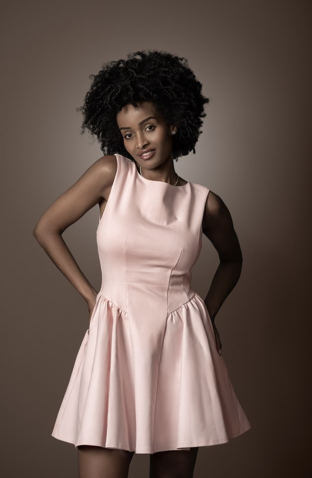 Mimi in a pale pink fit-and-flare dress, hands on hips, warm studio backdrop